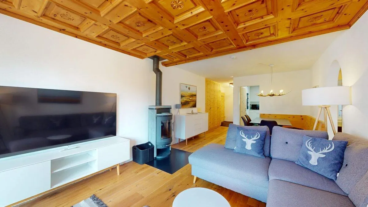 Wohnzimmer mit Fernseher, Ecksofa und Ofen unter der geschnitzten Holzdecke