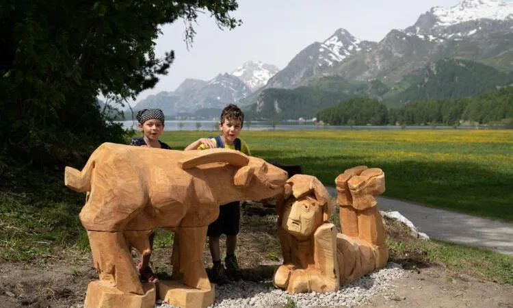 Zwei Kinder bei geschnitzten Holztieren am Märchenweg in Sils, im Hintergrund See und Berge