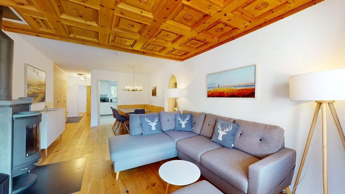 Wohnzimmer mit grauem Ecksofa, Arvenholzdecke und Specksteinofen, im Hintergrund der Essbereich